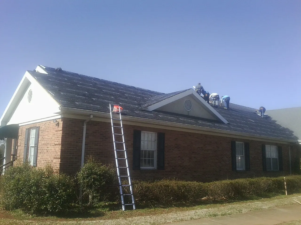 Crew tearing shingles off a brick ranch roof with a ladder set — Metal Roof Repair work by our crew in Clark Mills