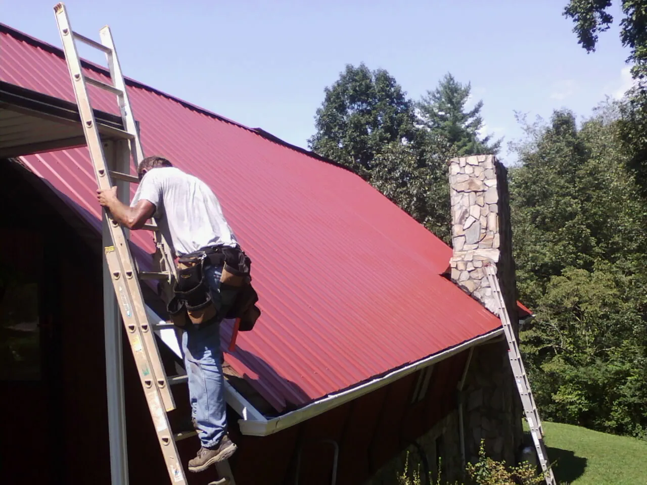 Roofer on a ladder beside a red metal roof and stone chimney — Soffit & Fascia Repair by Roofing Brothers in Clark Mills